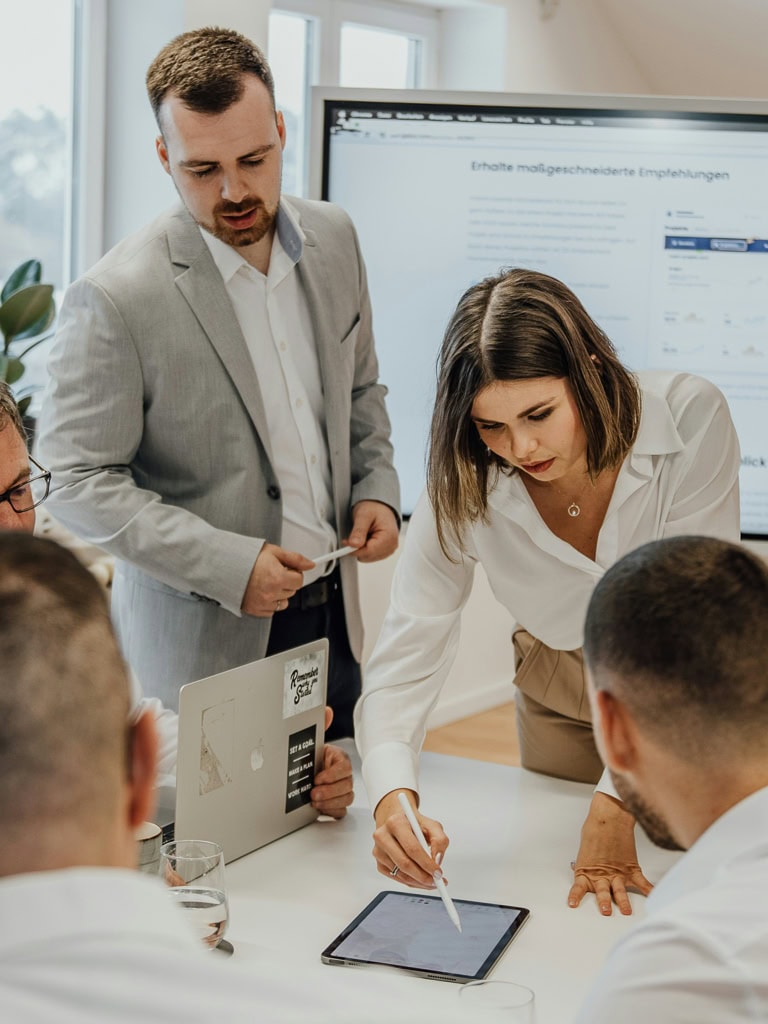 Un homme et une femme dans un bureau travaillent, devant eux un groupe d'employés et derrière eux un grand écran.